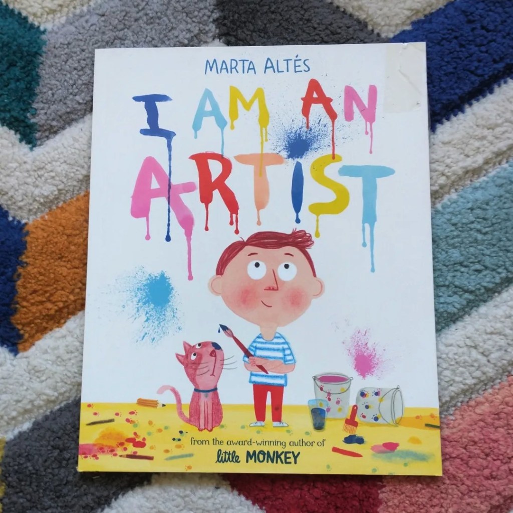 Marta Altes I Am An Artist Book Cover against a colorful rug. The cover shows a small boy surrounded by paint which has splashed on the floor, while crayons lie all around. His pet cat looks at a drop of paint which is about to fall from his brush while the boy looks proudly at the title I AM AN ARTIST that he appears to have painted on the wall.